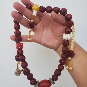 Peut inclure: Un collier de perles avec des perles rouge foncé sculptées avec des visages de type crâne, un dé rouge et un charme doré. Le collier comprend une grosse perle ovale rouge et une perle blanche et bulbeuse. Les perles sont enfilées sur un cordon jaune.