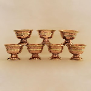 Puede incluir: Juego de seis cuencos de cobre con detalles dorados. Los cuencos tienen un acabado martillado y diseños florales intrincados. Están dispuestos en forma de pirámide sobre un fondo blanco.
