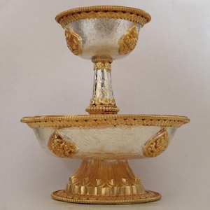 May include: A silver and gold tiered offering bowl with intricate floral designs. The bowl is made of metal and has a traditional Tibetan style.