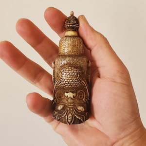 May include: A small, intricately carved, ivory-colored bottle stopper with a detailed design of a mythical creature's face. The stopper is topped with a small, ornate finial.
