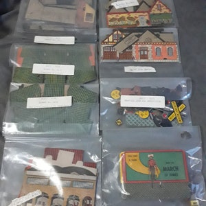 May include: Eight clear plastic bags containing various paper model kits. The kits include a schoolhouse, a fire station, a house, a bank, a traffic light, and a sign that says "Airport Crossing". There is also a small cardboard box with a picture of a cowboy and the text "March of Dimes".
