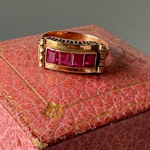 Peut inclure: Bague en or ornée de trois pierres rouges rectangulaires. La bague présente un design rectangulaire avec des détails décoratifs sur les côtés et le dessus. Elle est présentée sur un écrin à bijoux rose texturé.