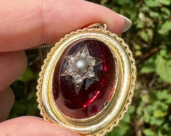 Könnte beinhalten: Goldfarbener Anhänger mit einem ovalen, tiefroten Mittelteil. Ein sternförmiges Design mit einem Perlzentrum und umliegenden klaren Steinen ist in der roten Mitte eingesetzt. Der Anhänger wird von einer Hand vor grünem Hintergrund gehalten.
