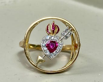 May include: A gold ring with a red ruby heart pierced by an arrow and surrounded by smaller diamonds. The heart is set in a circle with a flame design above it.