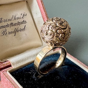 May include: A gold ring featuring a decorative sphere, set in a pink and blue velvet jewellery box. The sphere is covered in intricate scrollwork. The box is open, revealing the ring's band.