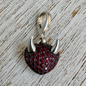 May include: A silver-toned heart-shaped pendant with a bail. The heart is encrusted with small, round, red gemstones. Two silver horns extend from the top of the heart.