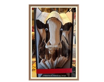 May include: A folded book art sculpture of a cow's face. The sculpture is made from the pages of a book and is painted in black and white. The sculpture is framed in a brown frame with the text "Folded Book Patterns by Jacqui" and "A45" on the bottom.