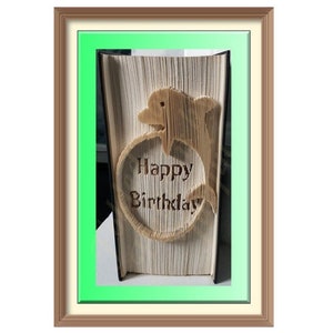 May include: A framed, open book with pages folded to create a dolphin design and a heart-shaped message that reads "Happy Birthday." The frame has a brown border with a green and cream-colored inner border. The book is the focal point.