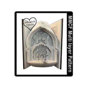 May include: A book with pages folded into a detailed nativity scene. The book is open, revealing the artwork, with the text "JHBookFold Patterns" in a heart-shaped logo. The image is framed with a black border and the text "MMCF Multi layer Pattern".