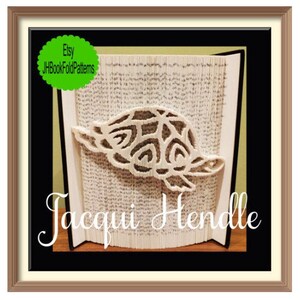 May include: A framed artwork featuring a turtle design cut from the pages of a book. The turtle is light-colored and detailed. The artwork is set against a backdrop of book pages, with the artist's name, "Jacqui Hendle," displayed below.