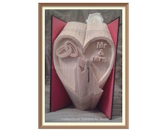 May include: A book folded into the shape of a heart with two wedding rings and the words "Mr & Mrs" on it. The heart is surrounded by red paper.