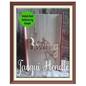 May include: A framed folded book art piece with a boxing theme. The word "Boxing" is cut out of the pages, along with a silhouette of a boxer. The artist's name, Jacqui Hendle, is written below.