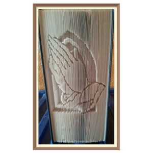 Puede incluir: Una escultura de arte de libro de un par de manos en posición de oración. La escultura está hecha de las páginas de un libro que se han doblado y cortado cuidadosamente para crear el diseño. La escultura se muestra en un marco marrón.