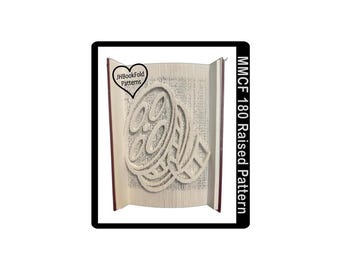 May include: A book folded into a 3D design of a film reel. The book is open and the design is visible. The text "JHBookFold Patterns" is visible in a heart shape on the book cover. The text "MMCF 180 Raised Pattern" is visible on the right side of the image.