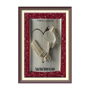 May include: A framed book art piece featuring a heart shape created from the pages of the book. The heart is adorned with a pair of scissors and a comb, hinting at a hairdressing theme. The frame is brown with a red glitter border. The text "Folded by LJ Book Art" and "Folded Book Patterns by Jacqui" is visible on the book.