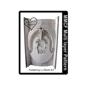 May include: A book art piece featuring a layered nativity scene cut into the pages. The book is folded to create a 3D effect, with the text "JHBookFold Patterns" and "MMCF Multi layer Pattern" visible. The piece is framed in black.
