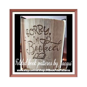Könnte beinhalten: Ein gefaltetes Buchkunst-Muster mit dem Text "Sorry, ich bin ausgebucht" und einem kleinen Buchsymbol. Das Muster wird durch Falten der Seiten eines Buches erstellt. Der Text ist in einer dekorativen Schriftart. Das Bild ist in einem braunen Rahmen gerahmt. Der Text "Gefaltete Buchmuster von Jacqui" und eine Website-Adresse befinden sich am unteren Rand des Bildes.