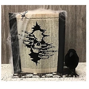 Pode incluir: Uma escultura de arte de livro em preto e branco de uma caveira com mãos que saem de um buraco no livro. O livro está rodeado de teias de aranha pretas e um corvo preto está sentado em uma prateleira abaixo do livro.