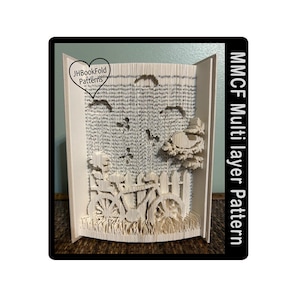 May include: A white book folded into a 3D sculpture of a bicycle with a fence and a bird's nest in a tree. The book is titled "MMCF Multilayer Pattern" and has a heart-shaped logo that says "JHBookFold Patterns".