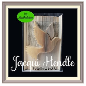 May include: A book art sculpture of a dove in flight, folded from the pages of a book. The sculpture is framed in a silver frame with a black mount. The artist's name, Jacqui Hendle, is written below the sculpture, along with the text "Folded by LJ Book Art".