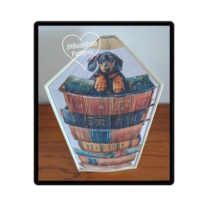 May include: A book folding art piece featuring a black and tan dachshund puppy peeking over the top of a stack of books. The books are arranged in a vertical stack and are all different sizes and colors. The puppy is looking directly at the viewer. The art piece is titled "JHBookFold Patterns".