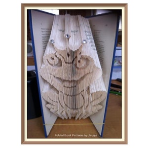 May include: A book folded into the shape of a frog. The frog is made of white paper and has a detailed design. The book is open and the frog is standing upright. The text "Folded Book Patterns by Jacqpa" is visible at the bottom of the image.