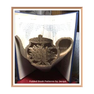 Può includere: Un libro piegato a forma di teiera con dettagli floreali. Il libro è aperto e le pagine sono piegate per creare la forma della teiera. Il testo "Folded Book Patterns by Jacqui" è visibile nella parte inferiore dell'immagine.