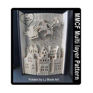 May include: A 3D book folding art piece depicting a Santa Claus flying over a town with houses. The text "Book Folding Patterns" is in a heart shape above Santa. The text "MMCF Multi layer Pattern" is on the right side of the image. The text "Folded by LJ Book Art" is at the bottom of the image.