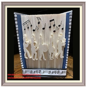 May include: A book art sculpture of a marching band with musical notes above them. The book is white with blue and black striped paper on the sides. The book is standing upright on a wooden surface.