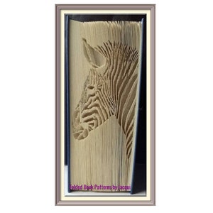 May include: A book art sculpture of a zebra's head, created by folding the pages of a book. The sculpture is framed in a light brown frame. The text "Folded Book Patterns by Jacqui" is visible at the bottom of the image.
