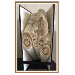 Könnte beinhalten: Ein Buch, das zu der Form einer Person gefaltet ist, die ein Dirtbike fährt. Das Buch ist geöffnet und die Seiten sind gefaltet, um die Form des Bikes und des Fahrers zu bilden. Das Buch befindet sich in einem schwarzen Rahmen.