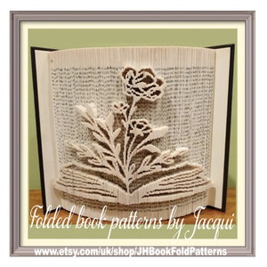 Può includere: Una scultura di arte libraria di un fiore con foglie e steli, piegata dalle pagine di un libro. Il libro è aperto e la scultura è in piedi. La scultura è incorniciata in una cornice marrone chiaro. Il testo "Folded book patterns by Jacqui" e "www.etsy.com/uk/shop/JHBookFoldPatterns" è visibile nella parte inferiore dell'immagine.