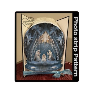 May include: A blue and white photo strip pattern for a bookfold craft project depicting a nativity scene with a star above the manger. The pattern is titled "Photo strip pattern" and includes the text "JHBookFold Patterns".
