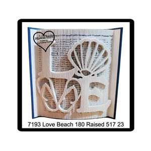 May include: A book with pages folded to create the word "LOVE" and a seashell design. The book is white with blue edges and the text "7193 Love Beach 180 Raised 517 23" at the bottom. A heart-shaped logo is in the upper left corner.