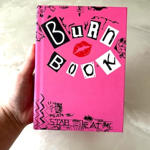 以下が含まれることがあります： 「Burn Book」というタイトルのピンク色の本。赤い唇のグラフィックを特徴とするコラージュデザイン。本のテキストとイラストは白黒です。本は手で持たれています。