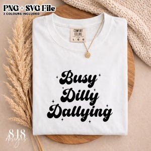 Op de afbeelding: Een wit t-shirt met de tekst "Busy Dilly Dallying" in zwart schrift. Het shirt heeft een ronde hals en een klein label met de tekst "Comfort Colors". Een gouden ketting met een zonvormige hanger ligt op het shirt. De woorden "PNG + SVG File" en "3 COLOURS INCLUDED" staan bovenaan.