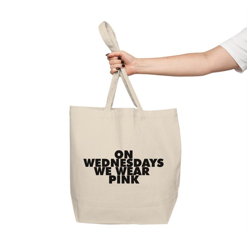 Może przedstawiać: Beżowa torba typu tote z czarnym napisem "ON WEDNESDAYS WE WEAR PINK". Torba ma dwa uchwyty i jest trzymana przez dłoń.