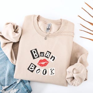 Może przedstawiać: Beżowa bluza z okrągłym dekoltem z napisem "Burn Book" w kolorze czarnym, czerwonym i białym. Projekt zawiera parę czerwonych ust. Srebrny naszyjnik jest zarzucony na bluzę. Bluza jest złożona na białej powierzchni.