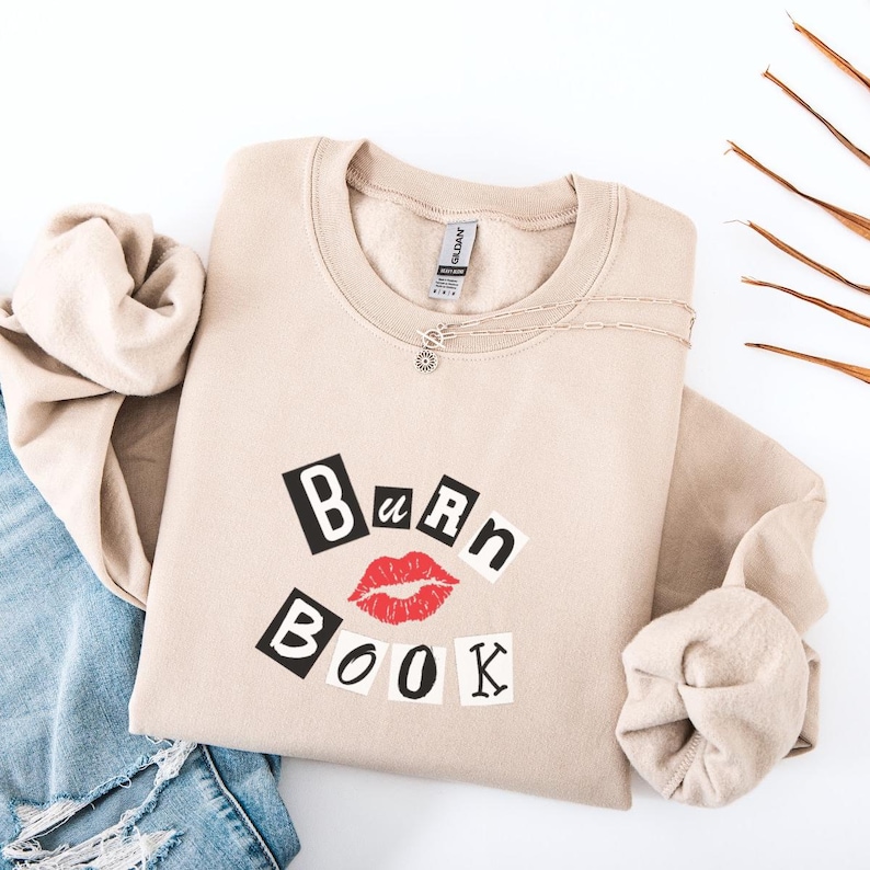 Może przedstawiać: Beżowa bluza z okrągłym dekoltem z napisem "Burn Book" w kolorze czarnym, czerwonym i białym. Projekt zawiera parę czerwonych ust. Srebrny naszyjnik jest zarzucony na bluzę. Bluza jest złożona na białej powierzchni.
