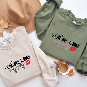 Może przedstawiać: Dwie bluzy, jedna beżowa i jedna oliwkowa, z napisem "You're Like Really Pretty" wydrukowanym w kolorze czarnym, r&oacute;żowym i czerwonym. Obok tekstu znajduje się grafika czerwonych ust. Na zdjęciu znajduje się r&oacute;wnież brązowy kapelusz i filiżanka kawy.