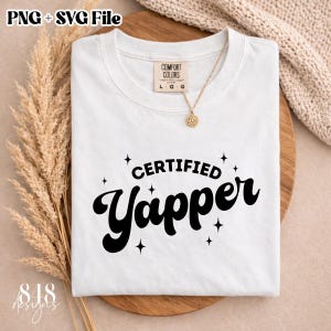 Op de afbeelding: Wit T-shirt met de zwarte tekst "CERTIFIED Yapper", met steraccenten. Het shirt heeft een ronde hals en een Comfort Colors-label. Een gouden ketting met een zonnehanger ligt op het shirt.