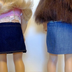 K&ouml;nnte beinhalten: Zwei Puppenr&ouml;cke: einer aus dunkelblauem Denim mit goldfarbenen N&auml;hten und einer aus hellblauem Denim. Die R&ouml;cke sind von hinten zu sehen, wobei die Beine der Puppen sichtbar sind. Der dunkelblaue Rock hat einen geraden Saum, der hellblaue Rock einen elastischen Bund.