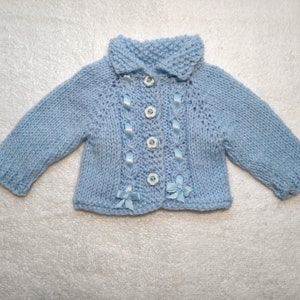 Puede incluir: Un c&aacute;rdigan de punto azul claro para beb&eacute; con cuello y botones blancos. El c&aacute;rdigan tiene detalles de cinta azul y botones blancos.