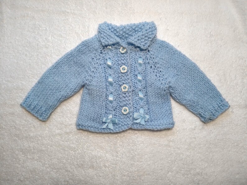 Puede incluir: Un c&aacute;rdigan de punto azul claro para beb&eacute; con cuello y botones blancos. El c&aacute;rdigan tiene detalles de cinta azul y botones blancos.