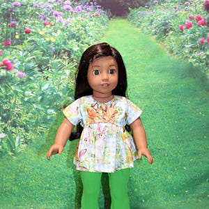 May include: A doll wearing a green and white dress with a bunny print and green pants. The doll has long brown hair and brown eyes. The doll is standing in a garden setting.