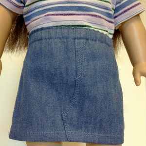 K&ouml;nnte beinhalten: Eine Puppe tr&auml;gt einen blauen Jeansrock und ein gestreiftes T-Shirt. Das T-Shirt hat horizontale Streifen in Blau-, Lila- und Gr&uuml;nt&ouml;nen. Der Rock ist im klassischen A-Linien-Stil gehalten.