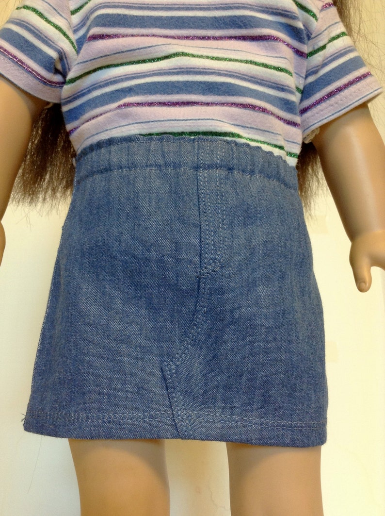 K&ouml;nnte beinhalten: Eine Puppe tr&auml;gt einen blauen Jeansrock und ein gestreiftes T-Shirt. Das T-Shirt hat horizontale Streifen in Blau-, Lila- und Gr&uuml;nt&ouml;nen. Der Rock ist im klassischen A-Linien-Stil gehalten.