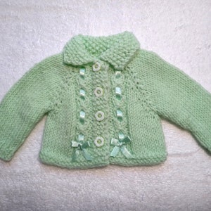 Puede incluir: Un c&aacute;rdigan de punto para beb&eacute; de color verde claro con cuello blanco y botones. El c&aacute;rdigan tiene botones blancos y detalles de cinta verde.