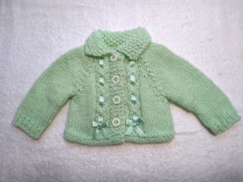 Puede incluir: Un c&aacute;rdigan de punto para beb&eacute; de color verde claro con cuello blanco y botones. El c&aacute;rdigan tiene botones blancos y detalles de cinta verde.