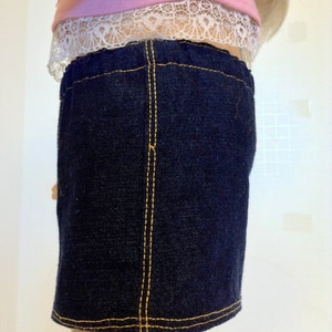 K&ouml;nnte beinhalten: Ein dunkelblauer Jeansrock mit gelben N&auml;hten. Der Rock ist Teil eines Puppenoutfits und wird mit einem rosa Oberteil mit wei&szlig;er Spitzenborte kombiniert. Der Rock hat ein klassisches, schlichtes Design.